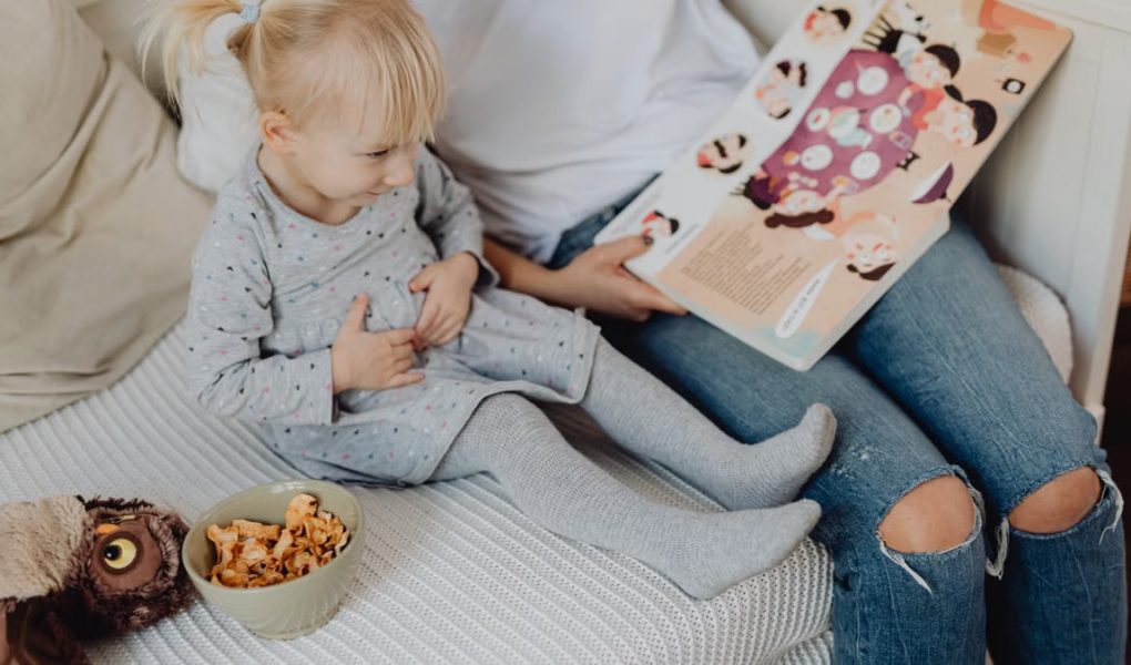 Mother reading a book to her daughter in a cozy bedroom setting with snacks nearby.