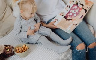 Mother reading a book to her daughter in a cozy bedroom setting with snacks nearby.