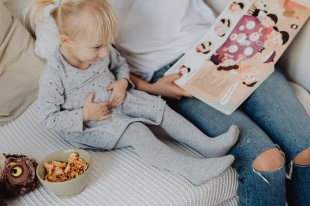 Mother reading a book to her daughter in a cozy bedroom setting with snacks nearby.