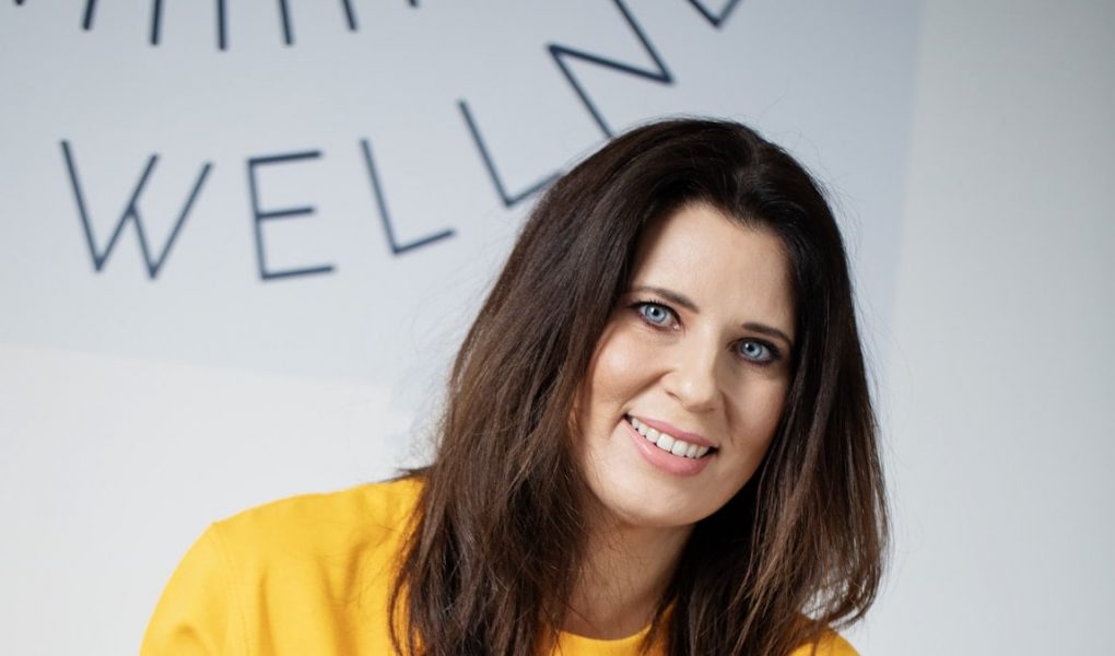A woman with dark hair smiles in a yellow sweatshirt.