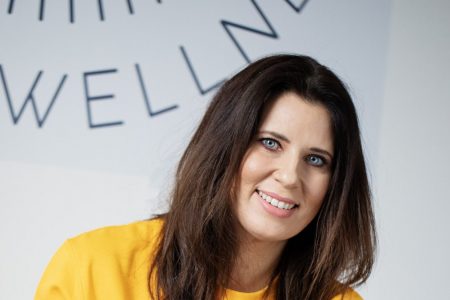 A woman with dark hair smiles in a yellow sweatshirt.